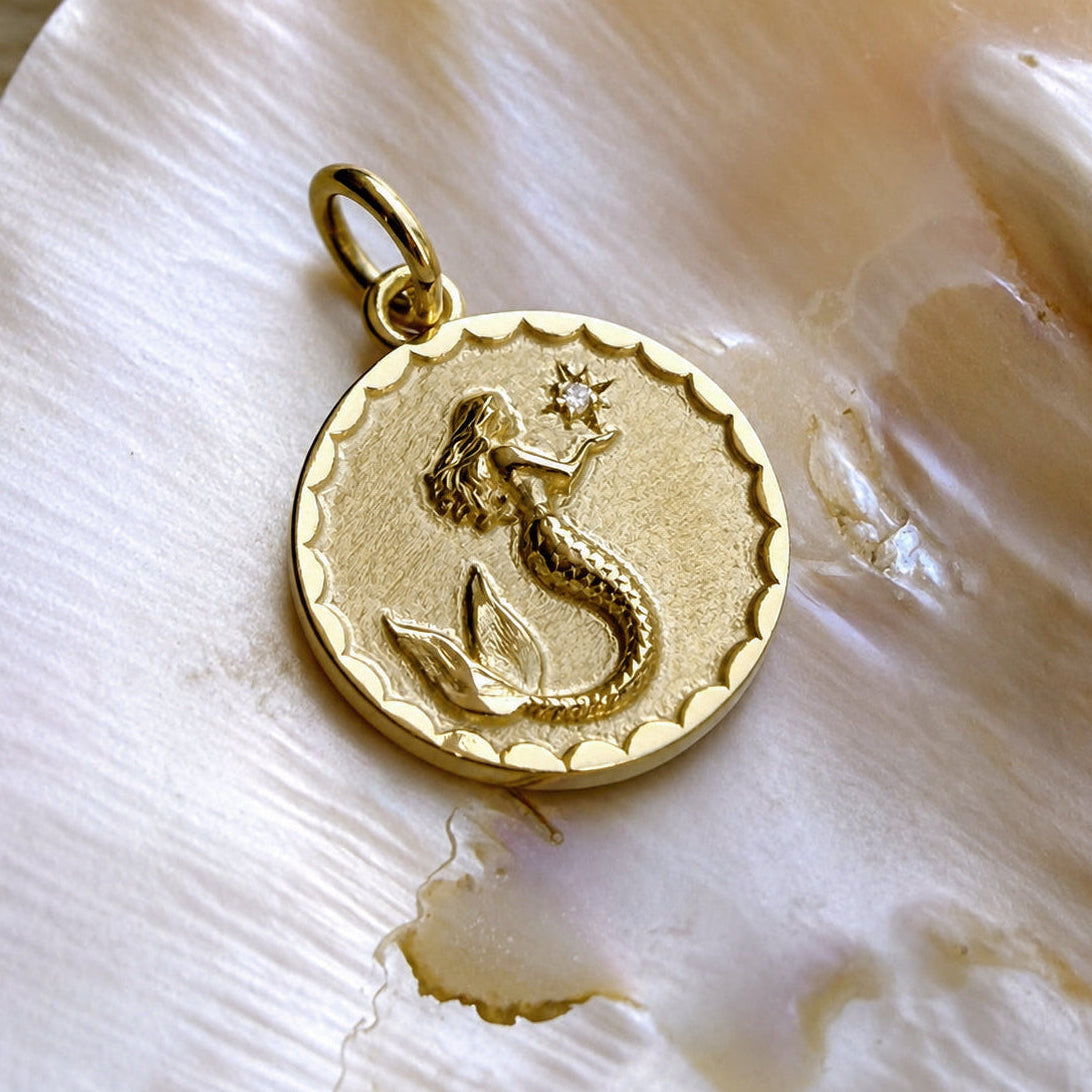 A gold circular pendant charm featuring an embossed mermaid holding a star, displayed on a white seashell.